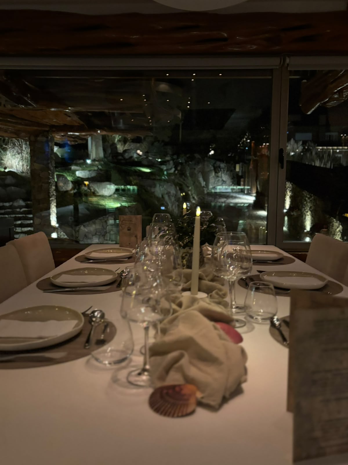 Mesa larga puesta para la cena, con velas y el jardín iluminado detrás del ventanal